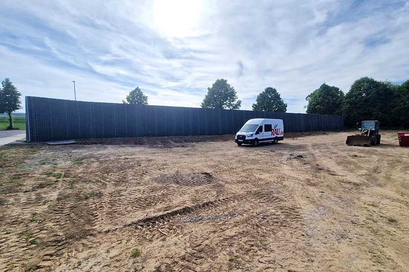 RAU climate wall made of grid elements on a sandy construction site. A white van with the RAU logo and a small wheel loader are parked in front of a row of trees.