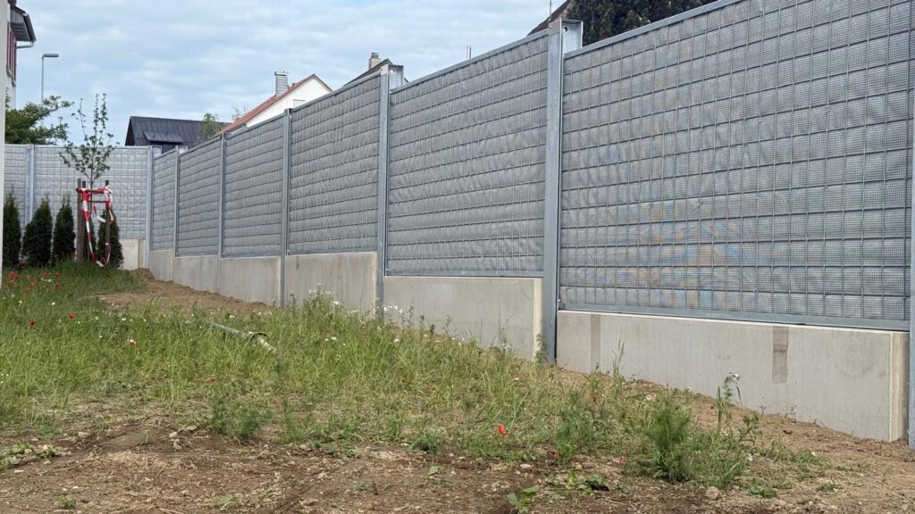 Eine hohe hellgraue Lärmschutzwand mit Metallgittern zieht sich entlang eines Grundstücks in einem Wohngebiet.