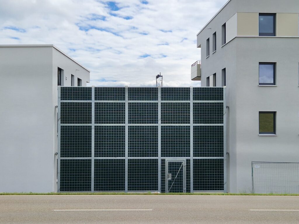 Un mur anti-bruit de grande taille, de couleur sombre, avec un remplissage de grilles métalliques, relie deux bâtiments résidentiels. Dans la partie inférieure droite du mur, une porte est encastrée et affleurante.