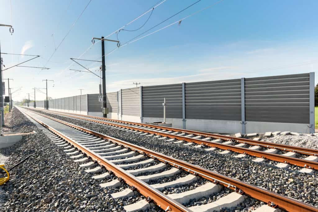 Bahngleise mit Oberleitungen verlaufen entlang einer hohen Schallschutzwand unter blauem Himmel