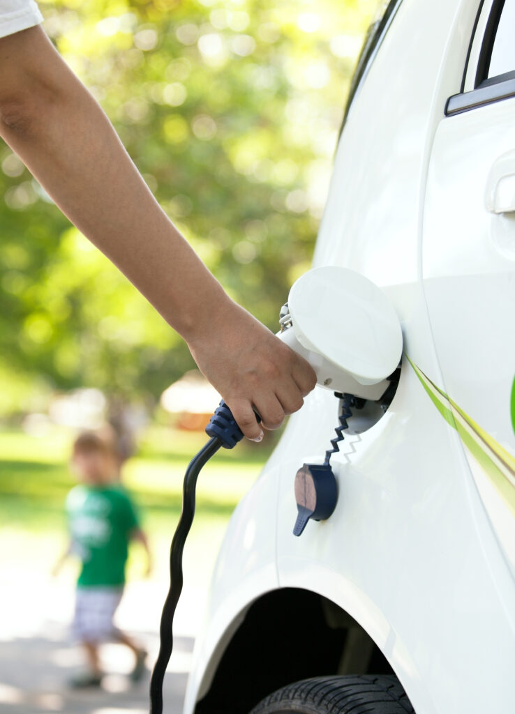 Eine Hand steckt ein Ladekabel in ein weißes Elektroauto, im Hintergrund ein spielendes Kind
