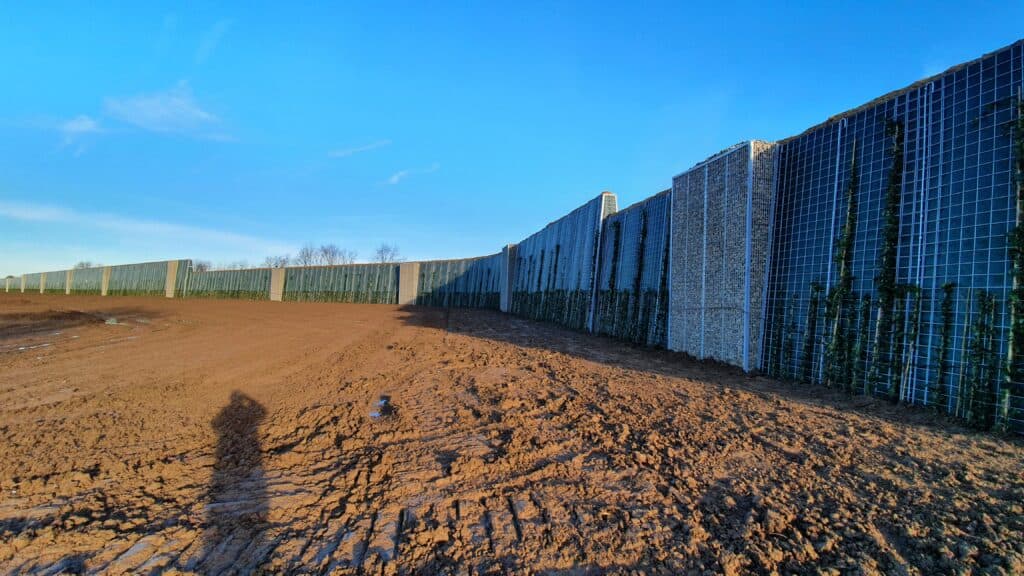 Lärmschutzwand aus Drahtgitterkörben entlang einer unbefestigten Fläche bei blauem Himmel