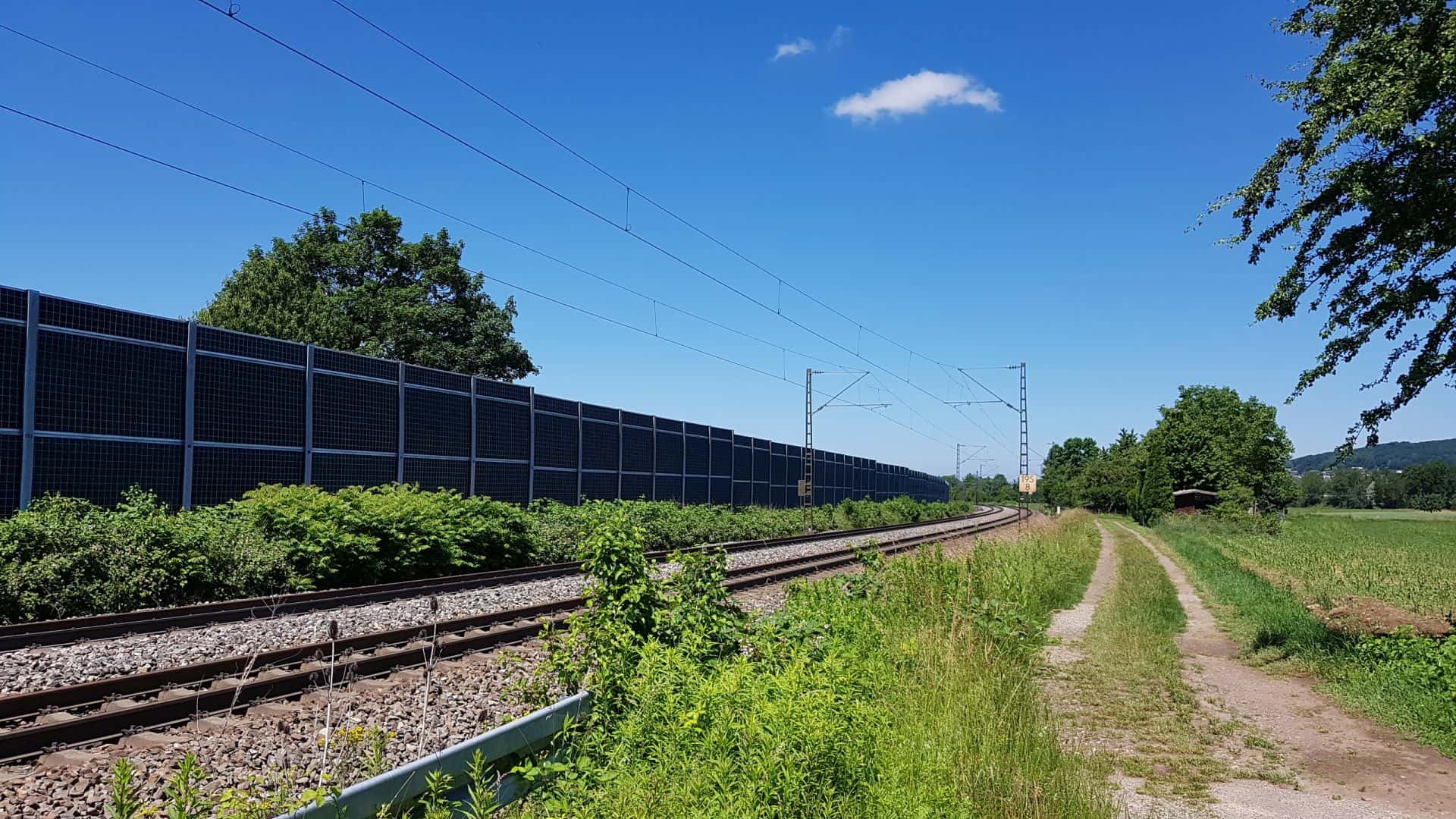 Lärmschutzwand entlang von Eisenbahngleisen und einem Feldweg bei sonnigem Wetter