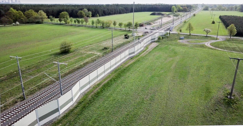 Luftaufnahme von Bahngleisen mit Lärmschutzwand, umgeben von grünen Feldern